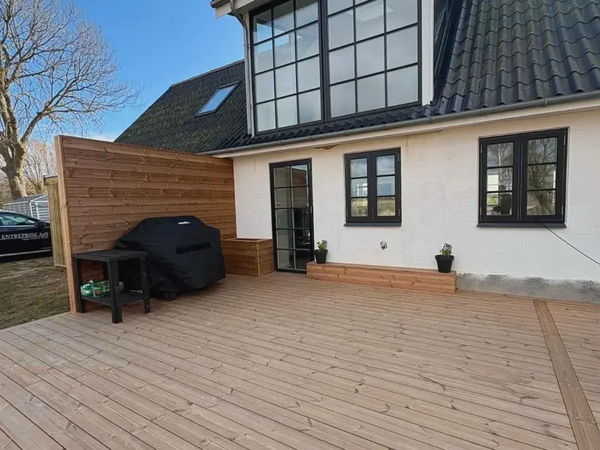 Træterrasse ved hvidt hus med sorte vinduespartier og læskærm